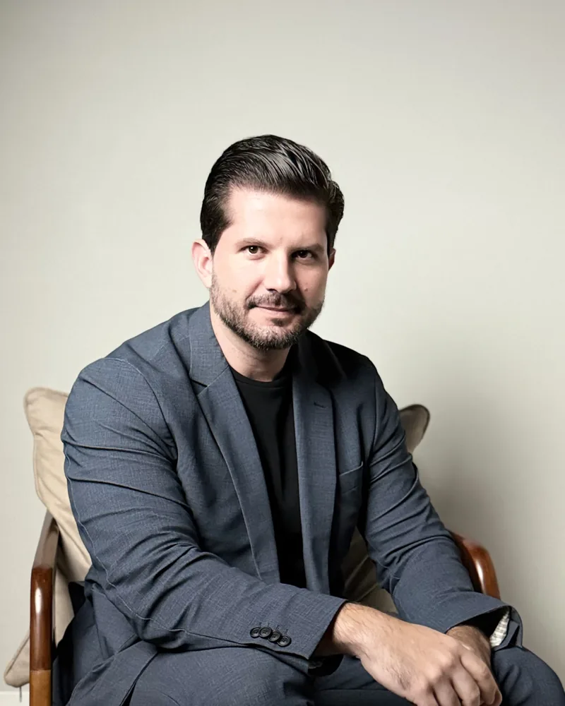 Dr. Joaquim Menezes sentado, de blazer cinza, com a mão apoiada no joelho